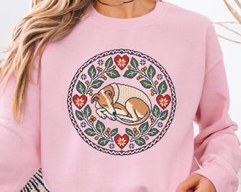 May include: A light pink crewneck sweatshirt featuring a circular graphic print of a sleeping tan and white dog curled up inside a wreath of green leaves, red hearts, and small yellow berries, with a decorative geometric border.