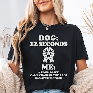May include: A black t-shirt featuring white text that reads DOG: 12 SECONDS, followed by a graphic of a circular award ribbon with a star in the center, and the text ME: 4 HOUR DRIVE, CAMP CHAIR IN THE RAIN, GAS STATION FOOD printed below.