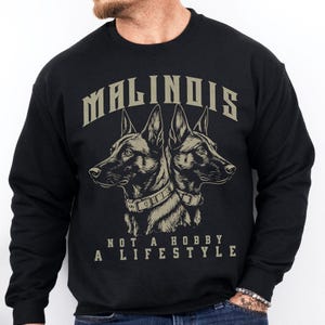 May include: A black long-sleeve sweatshirt featuring a graphic of two Belgian Malinois dogs facing opposite directions. Above the dogs is the text MALINOIS in a bold font, and below them is the text NOT A HOBBY A LIFESTYLE in a simple font.