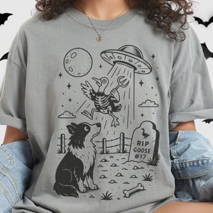 Puede incluir: Camiseta gris con un gráfico en blanco y negro. El gráfico muestra un OVNI abduciendo un ganso esqueleto, un perro mirando hacia arriba, una lápida que dice "RIP GOOSE #17", una luna y un hueso.