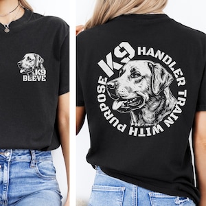 May include: Black t-shirt featuring a detailed illustration of a dog's face. The front of the shirt has the text "K9 BLEVE" and a dog graphic. The back of the shirt has the text "K9 Handler Train With Purpose" around a dog graphic.