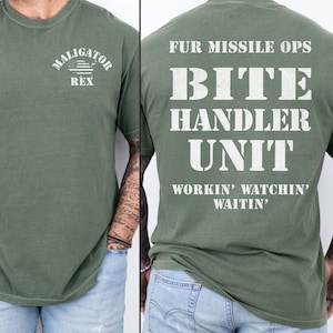 May include: Olive green t-shirt with white text and a graphic on the front that says "MALIGATOR REX" with an American flag design. The back of the shirt says "FUR MISSILE OPS BITE HANDLER UNIT WORKIN' WATCHIN' WAITIN'."
