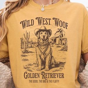 May include: Mustard-colored t-shirt with a graphic of a Golden Retriever wearing a cowboy hat and bandana. The text reads "Wild West Woof" and "Golden Retriever". Below the dog is the text "The Good, The Bad & The Fluffy".