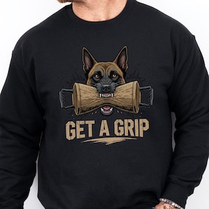 Puede incluir: Sudadera negra con un diseño de un perro pastor alemán sosteniendo una herramienta de entrenamiento de madera. El perro es marrón y negro con una expresión intensa. El texto "GET A GRIP" está impreso debajo en color beige.