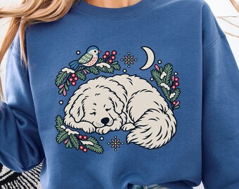 May include: A blue crewneck sweatshirt featuring a graphic print of a white fluffy dog sleeping curled up. The design includes green pine branches with red berries, a small blue bird, a crescent moon, and star-like floral accents.