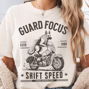 May include: An off-white t-shirt featuring a black graphic of a German Shepherd riding a motorcycle. The text reads GUARD FOCUS, ESTD 1989, SHIFT SPEED, and GERMAN SHEPHERD CREW. The design includes motion lines and a checkered pattern at the bottom.