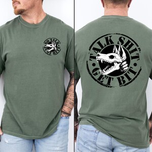 May include: An olive green t-shirt featuring a black and white dog skull graphic. The front displays the text "NO TALK JUST BITE" in a circular design. The back of the shirt has the text "TALK SHIT GET BIT" in a circular design.