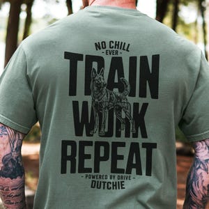 May include: Olive green t-shirt with the text "NO CHILL - EVER - TRAIN WORK REPEAT" in black. A detailed illustration of a dog is centered within the text. The text "- POWERED BY DRIVE - DUTCHIE" is at the bottom.