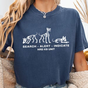 Puede incluir: Una camiseta azul marino con un gráfico blanco de tres esqueletos de perro en diferentes posturas. Debajo de los esqueletos, el texto dice SEARCH • ALERT • INDICATE y HRD K9 UNIT. La camiseta tiene cuello redondo y mangas cortas.
