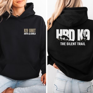 May include: A black hooded sweatshirt featuring a front graphic that reads K9 UNIT ARYA & KARLA. The back displays a large white graphic with the text HRD K9 THE SILENT TRAIL, accompanied by a silhouette of a dog walking through a wooded landscape.
