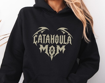 May include: A black hooded sweatshirt featuring a light beige graphic on the chest. The design is shaped like a heart with stylized wings at the top and the text "CATAHOULA MOM" printed in a bold, sharp, angular font in the center.