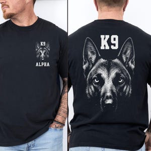 Puede incluir: Una camiseta negra de manga corta con un gráfico blanco de la cara de un pastor alemán. El pecho muestra el texto K9 ALPHA sobre el gráfico. La parte posterior de la camiseta presenta una versión grande del mismo gráfico con K9 arriba.