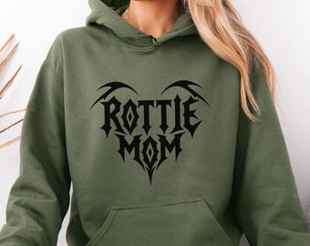 May include: An olive green hooded sweatshirt featuring a black graphic print on the chest. The design consists of the text ROTTIE MOM arranged in a heart shape with sharp, angular lettering and decorative pointed accents at the top and bottom.