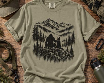 May include: A sage green short-sleeve t-shirt featuring a black graphic print of a large creature sitting on a rock with a dog, set against a backdrop of pine trees and mountains. The shirt has a crew neckline and a Comfort Colors brand label.