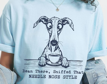 May include: Light blue t-shirt featuring a whimsical illustration of a long-nosed dog with a cup of coffee. The graphic includes the text "Bean There, Sniffed That NEEDLE NOSE STYLE" in a playful font. The shirt has short sleeves.