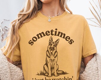 May include: A mustard yellow t-shirt featuring a dark brown graphic of a German Shepherd sitting in grass. The text above the dog reads "Sometimes" and the text below the dog reads "i just be watchin'" in a dark brown, rounded serif font.