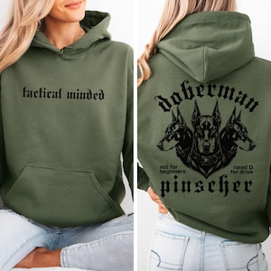 May include: An olive green hooded sweatshirt featuring the text tactical minded on the front. The back displays a graphic of three Doberman Pinscher heads with the text doberman pinscher, not for beginners, and rated D for drive in black ink.