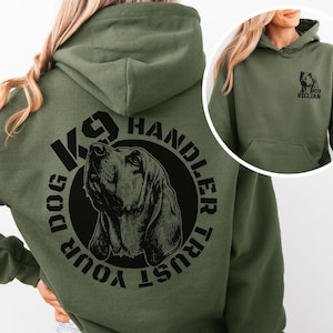May include: Olive green hoodie featuring a black graphic of a dog's head inside a circle with the text "TRUST YOUR DOG K9 HANDLER". The hoodie also has a small graphic of a dog and the text "K9 KILLIAN" on the front.
