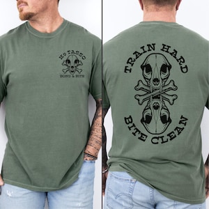 May include: An olive green short-sleeve t-shirt featuring a black graphic on the front left chest of a canine skull with crossed bones and the text K9 TASSO BOND & BITE. The back displays a larger version with the text TRAIN HARD BITE CLEAN.