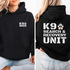Puede incluir: Una sudadera negra con capucha con texto blanco y un gráfico de huella. El frente muestra K9 BUCK con una pequeña huella. La parte posterior presenta un texto grande en blanco K9 SEARCH & RECOVERY UNIT con una huella que contiene una cruz naranja.