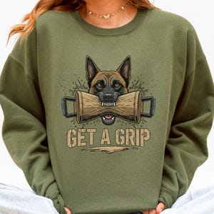 May include: Olive green sweatshirt featuring a graphic of a dog with a log in its mouth, with the text "GET A GRIP" in a tan color. The dog is brown and black with an intense expression. The sweatshirt is a casual style.