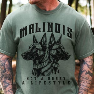 May include: A sage green short-sleeve t-shirt featuring a black graphic of two Belgian Malinois dogs facing opposite directions. The text MALINOIS is arched above the dogs, and the text NOT A HOBBY A LIFESTYLE is printed below the graphic.