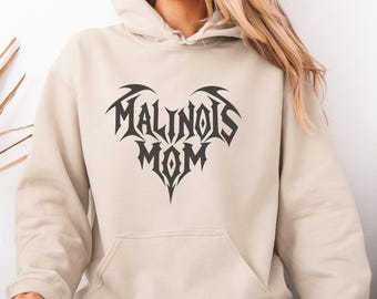 May include: A beige hooded sweatshirt featuring a dark gray graphic print on the chest. The design consists of the text MALINOIS MOM arranged in a heart shape with a sharp, angular, tribal-inspired font style. The hoodie includes a front pouch pocket.