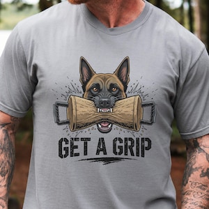 May include: A gray t-shirt featuring a detailed illustration of a German Shepherd dog with a wooden training tool in its mouth. The text "GET A GRIP" is printed below the dog in bold, black letters. The design has a distressed, vintage style.