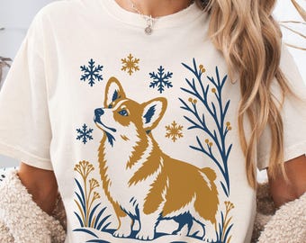 May include: A cream-colored short-sleeve t-shirt featuring a graphic print of a brown and white Corgi dog. The design includes dark blue and gold snowflakes, stylized branches, and floral elements arranged around the dog on a snowy ground.