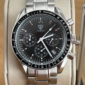 Puede incluir: Reloj cronógrafo Trident de acero inoxidable con esfera negra. El reloj presenta múltiples subesferas, una ventana de fecha y un bisel taquimétrico. Se muestra sobre un cojín color crema.