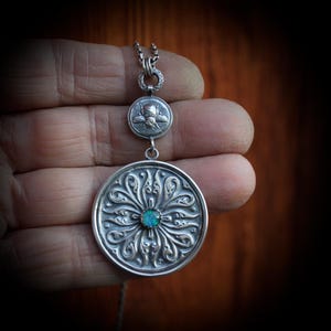 May include: A silver pendant necklace featuring a large, detailed circular pendant with an ornate floral design and a small, round turquoise stone in the center. The necklace also includes a smaller, round charm with a bee design.