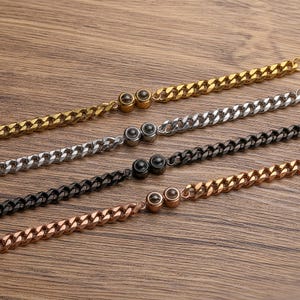 May include: Four curb chain bracelets in gold, silver, black, and rose gold. Each bracelet has a matching clasp with a small, round accent. The bracelets are displayed on a wooden surface.
