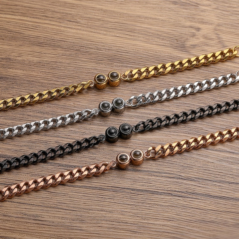 May include: Four curb chain bracelets in gold, silver, black, and rose gold. Each bracelet has a matching clasp with a small, round accent. The bracelets are displayed on a wooden surface.