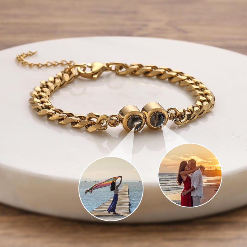 May include: Gold-colored chain bracelet with two round, photo-projecting charms. The bracelet is displayed on a white surface. Two small inset images show a woman on a pier and a couple on a beach.