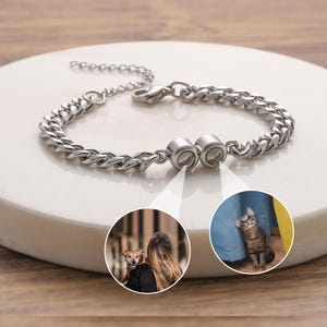 May include: A silver-colored bracelet with a curb chain design. The bracelet features two circular charms with images of a dog and a cat. The bracelet is displayed on a white surface.
