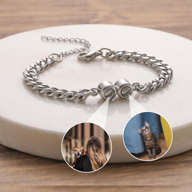 May include: A silver-colored bracelet with a curb chain design. The bracelet features two circular charms with images of a dog and a cat. The bracelet is displayed on a white surface.