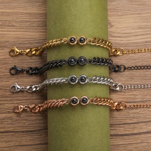 May include: Four bracelets in gold, black, silver, and rose gold, each with a curb chain design. Each bracelet features two small, dark, round beads. The bracelets are displayed on a green cylindrical object against a wooden background.