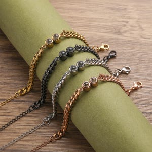 May include: Four chain bracelets in gold, black, silver, and rose gold. Each bracelet features a series of small, round, clear stones. The bracelets are displayed on a green, cylindrical surface against a wooden background.