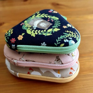 May include: Three fabric pouches stacked on top of each other. The top pouch is black with a green zipper and a floral pattern with a bear. The middle pouch is pink with a white zipper and a bird pattern. The bottom pouch is beige with a white zipper and a bird pattern.