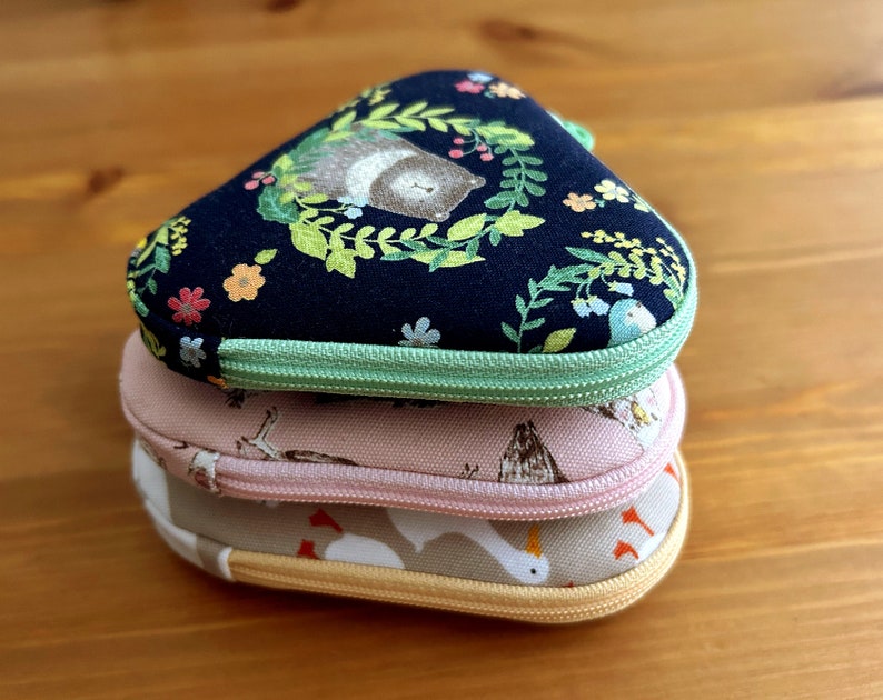 May include: Three fabric pouches stacked on top of each other. The top pouch is black with a green zipper and a floral pattern with a bear. The middle pouch is pink with a white zipper and a bird pattern. The bottom pouch is beige with a white zipper and a bird pattern.