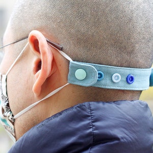 May include: A light blue fabric headband with buttons and a turquoise button on one end. The headband is worn by a person to hold a face mask in place.