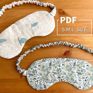 Könnte beinhalten: Zwei Augenmasken mit Blumenmustern und Gummibändern. Die Augenmasken sind aus weißem Stoff mit blauen und grünen Blumenmustern. Der Text "PDF S.M.L SIZE" ist auf einem weißen Rechteck gedruckt.