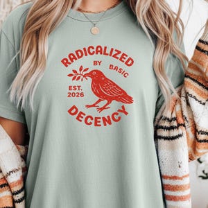 Puede incluir: Camiseta verde claro con la frase roja "RADICALIZED BY BASIC DECENCY" y un gráfico de pájaro rojo. La camiseta también muestra "EST. 2026". La camiseta está hecha de un material suave.