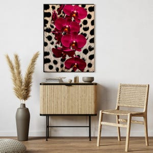 May include: A framed print of deep magenta orchids set against a cream-coloured background with black spots. The artwork is displayed above a wooden cabinet and a woven chair, creating a stylish interior.