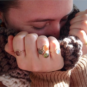 Op de afbeelding: Een persoon draagt een bruine gebreide sjaal en verschillende gouden ringen met gekleurde edelstenen. De ringen hebben verschillende stenen, waaronder groen, oranje en wit.