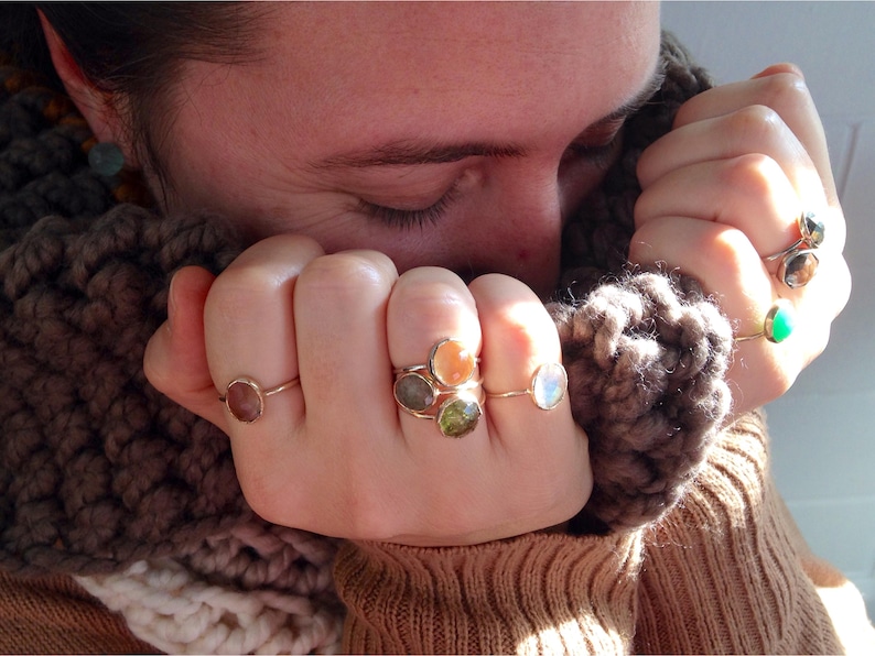 Op de afbeelding: Een persoon draagt een bruine gebreide sjaal en verschillende gouden ringen met gekleurde edelstenen. De ringen hebben verschillende stenen, waaronder groen, oranje en wit.