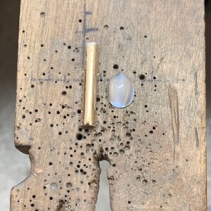 Op de afbeelding: Een close-up van een gouden metalen staaf en een witte ovale edelsteen op een houten oppervlak.