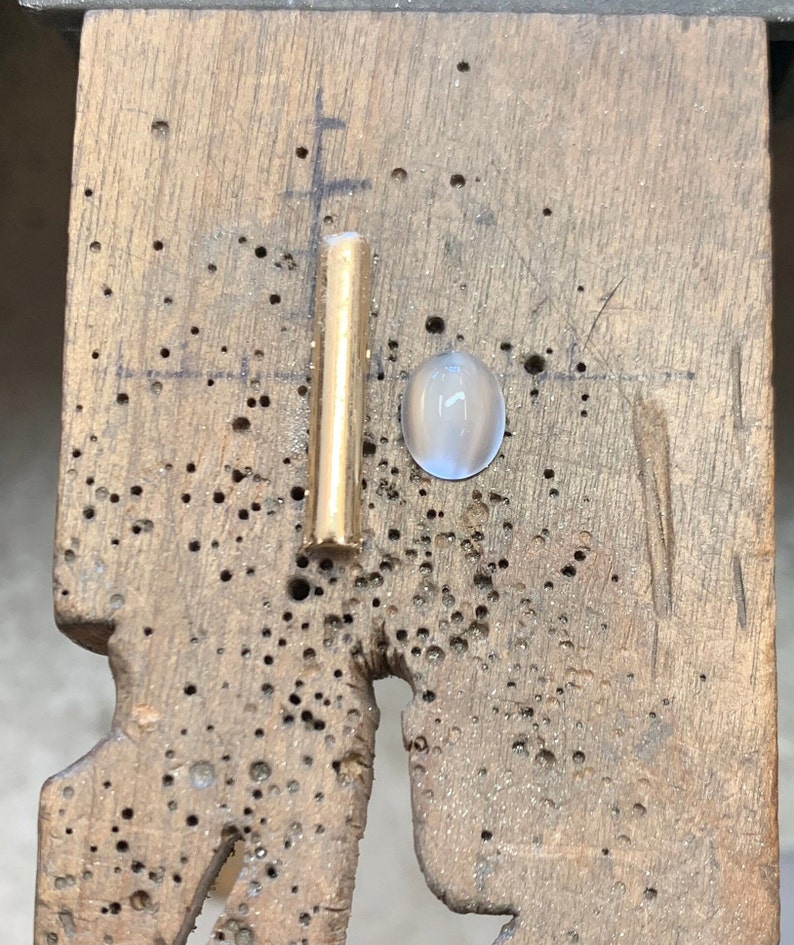 Op de afbeelding: Een close-up van een gouden metalen staaf en een witte ovale edelsteen op een houten oppervlak.