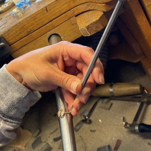 Op de afbeelding: Een hand gebruikt een metalen vijl om de randen van een gouden ring met een ronde stenen zetting glad te maken.