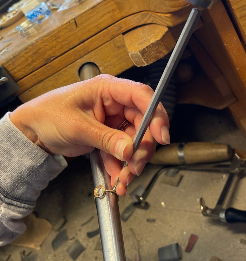 Op de afbeelding: Een hand gebruikt een metalen vijl om de randen van een gouden ring met een ronde stenen zetting glad te maken.